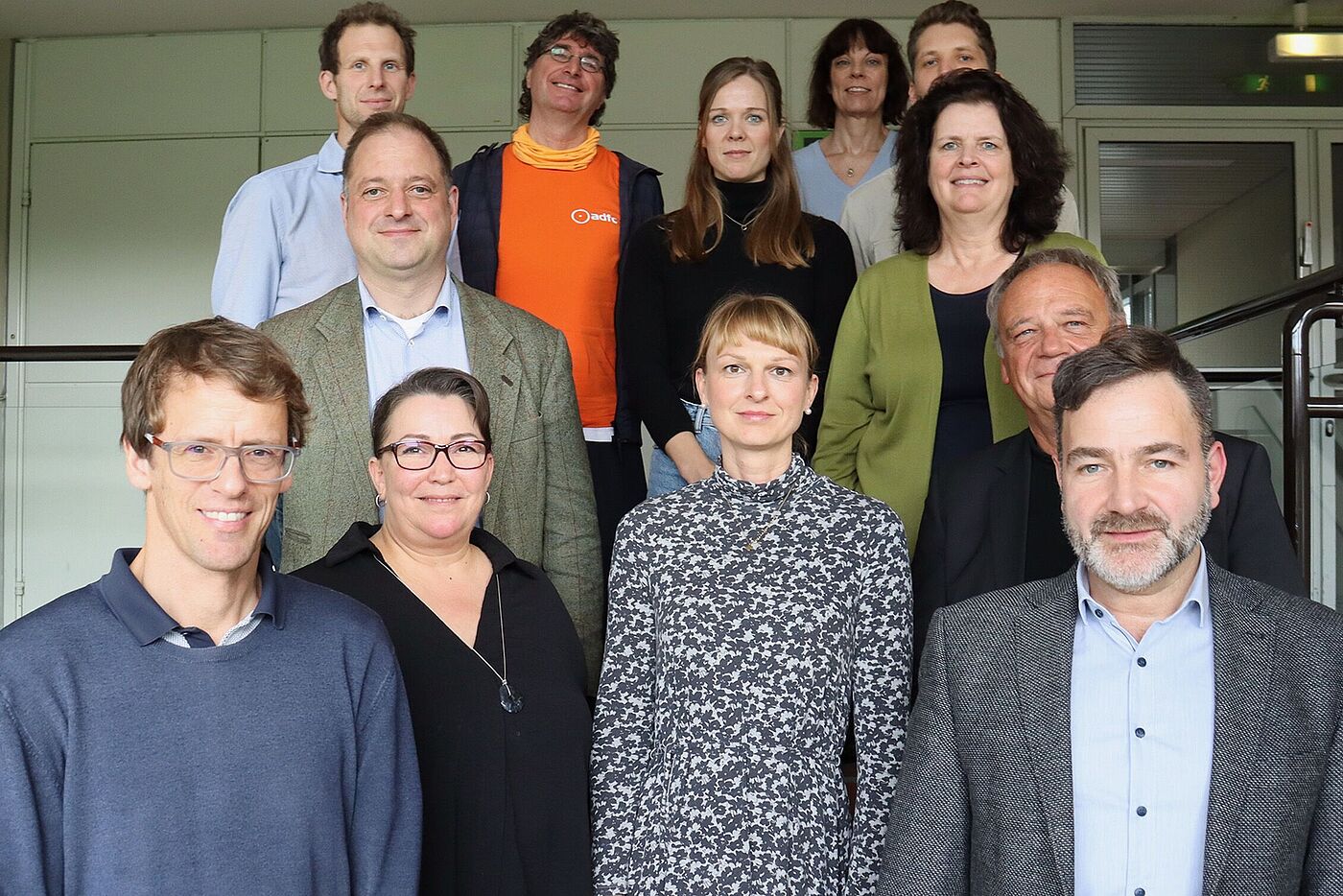 Zwölf Menschen stehen auf einem Podest. Man sieht nur ihre Oberkörper. Im Hintergrund ist eine Schrankwand zu erkennen.