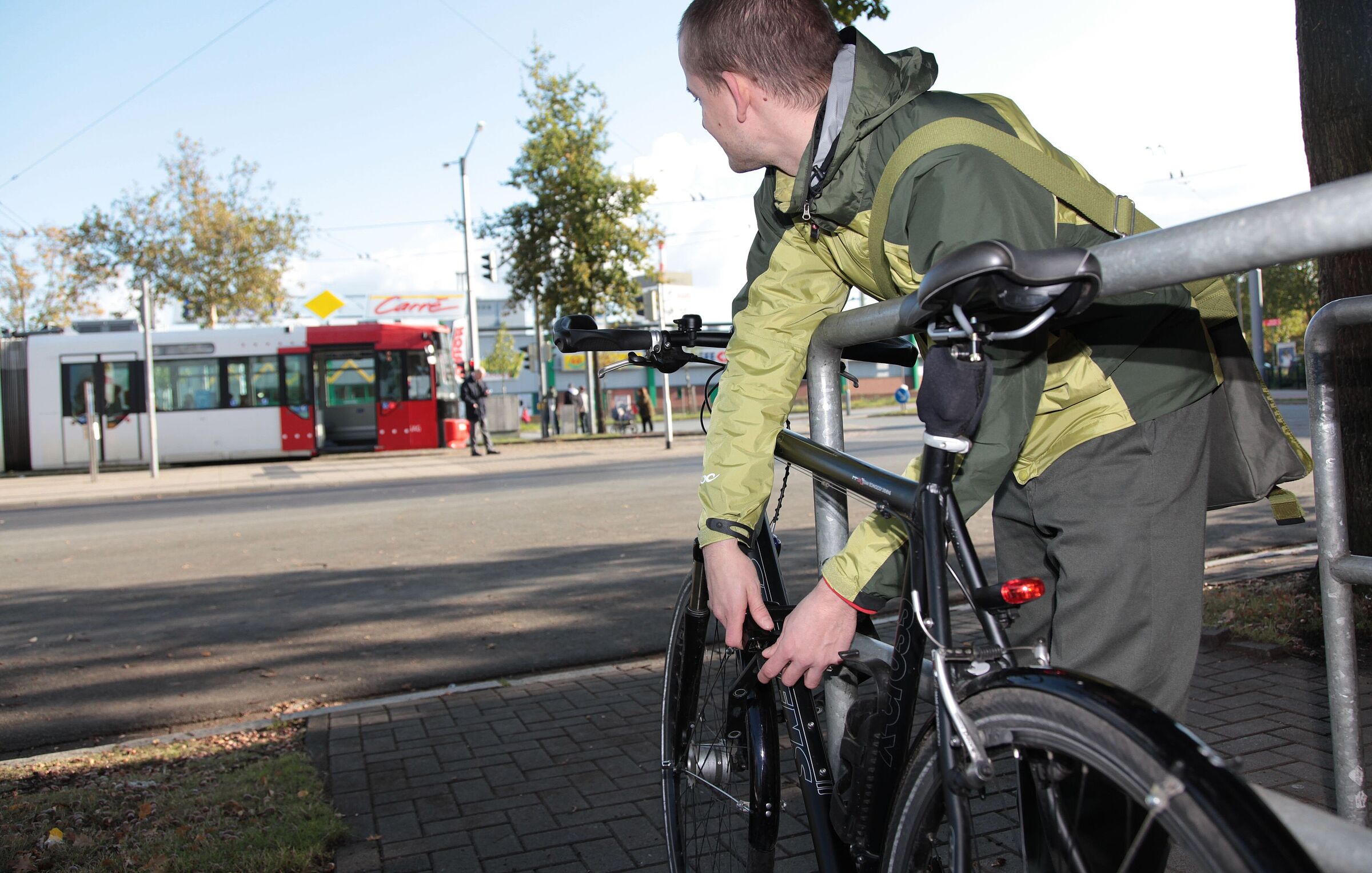 Ein junger mann schließt sein Fahrrad an einem Fahrradbügel an und schaut zu einer Straßenbahn im Hintergrund. 